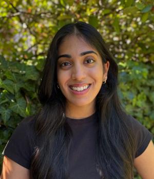 Person smilling with long dark hair and dark black shirt with light brown skin outside