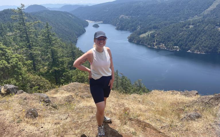 Photo of Emily Booker on top of a mountain overlooking a river. The river is surrounded by evergreen trees